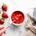 A hand dipping a spoon into a bowl of homemade strawberry sauce.