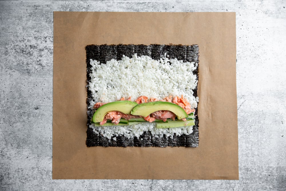salmon and avocado atop the cucumbers and rice on a sheet of nori for spicy salmon rolls