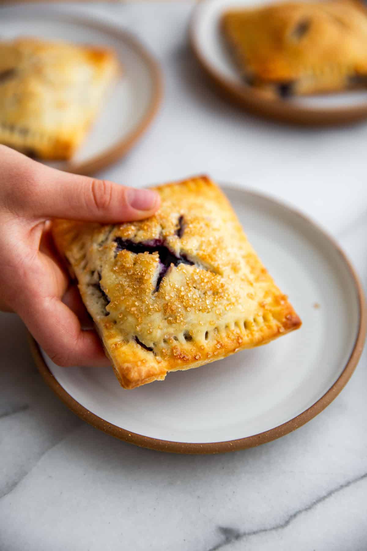 a hand holding a blueberry hand pie over a white plate