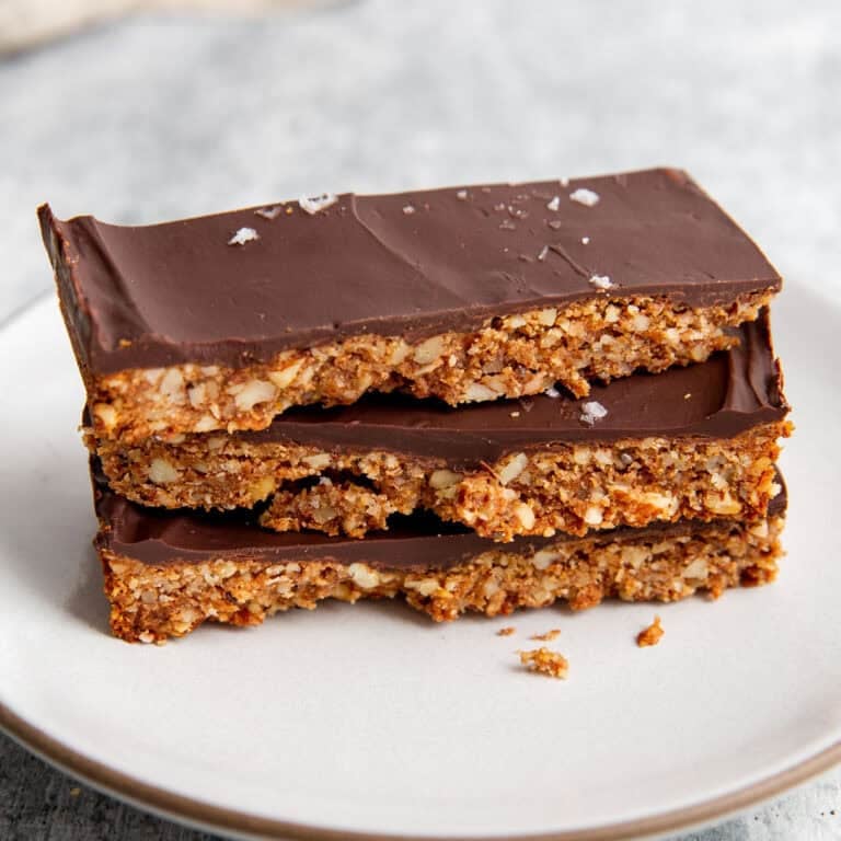 Three stacked chocolate-covered grain-free granola bars on a white plate. The bars have a crunchy texture with visible nuts and are topped with a layer of smooth, dark chocolate. A few flakes of sea salt are sprinkled on top.