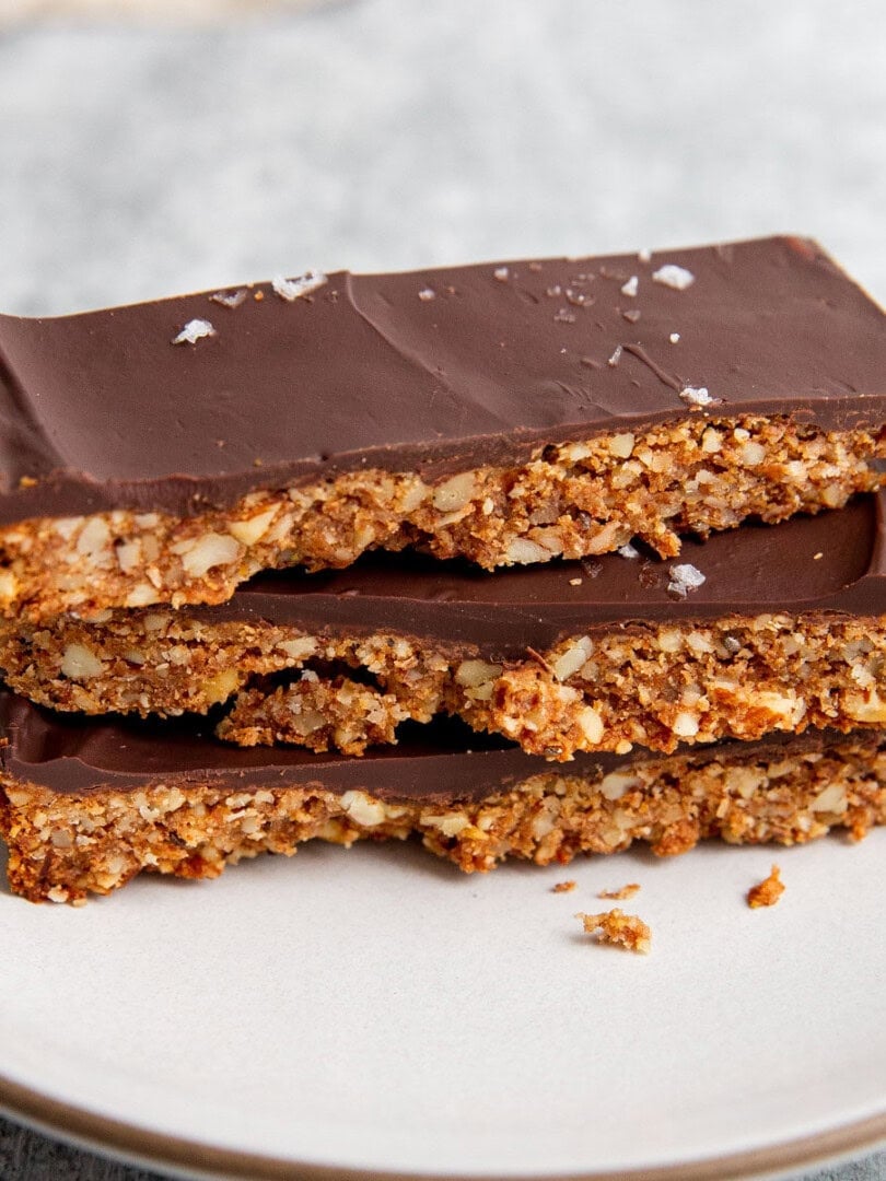 Three stacked chocolate-covered grain-free granola bars on a white plate. The bars have a crunchy texture with visible nuts and are topped with a layer of smooth, dark chocolate. A few flakes of sea salt are sprinkled on top.