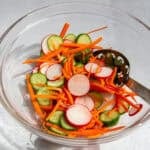 shredded carrots, sliced cucumbers, and sliced radishes in a glass bowl and a metal spoon for quick pickled vegetables.