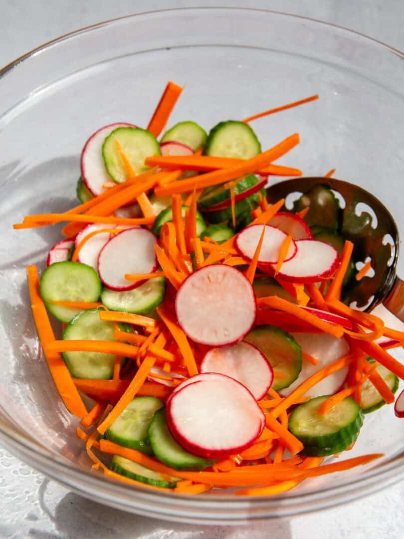 shredded carrots, sliced cucumbers, and sliced radishes in a glass bowl and a metal spoon for quick pickled vegetables.
