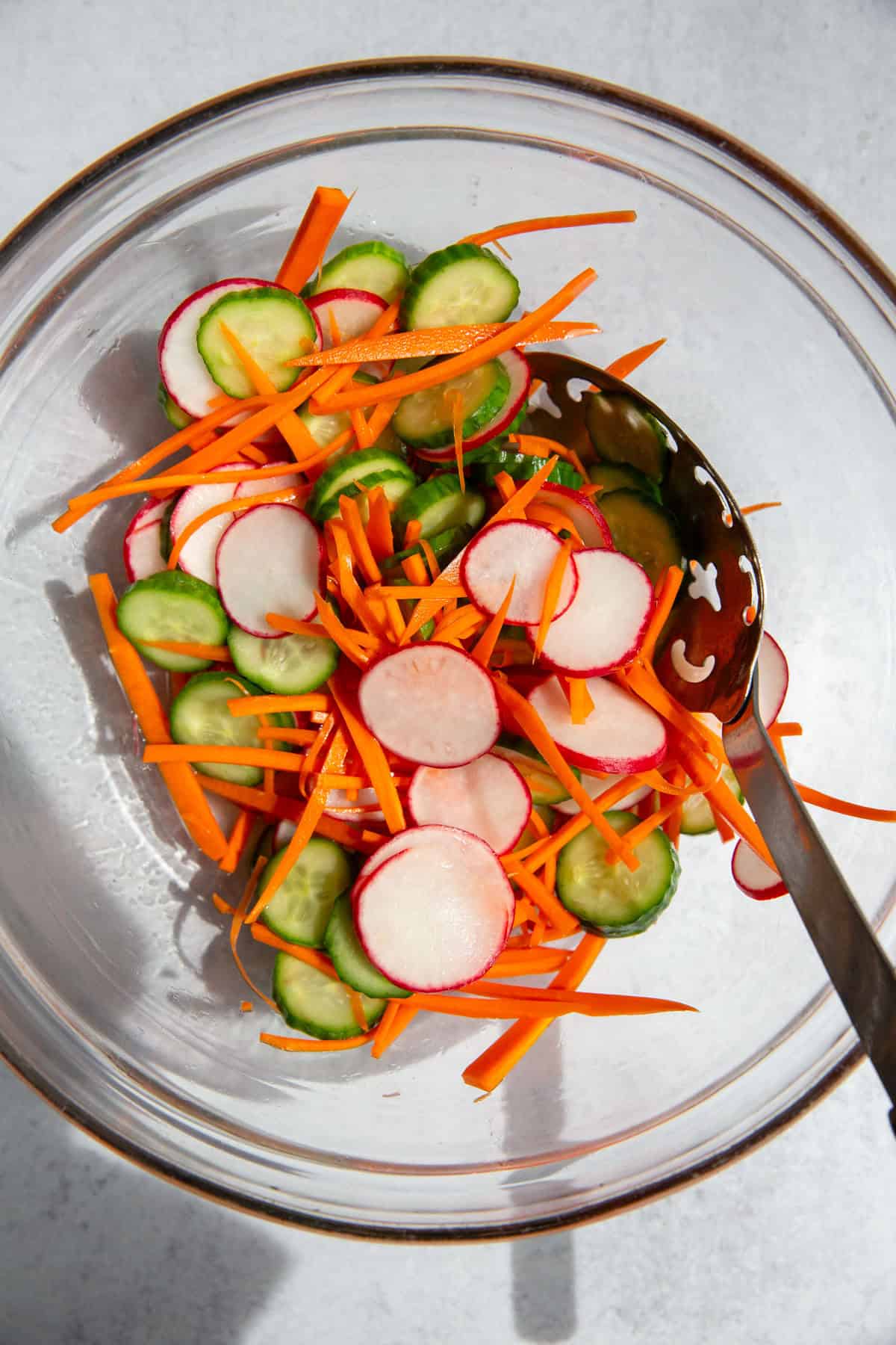shredded carrots, sliced cucumbers, and sliced radishes in a glass bowl and a metal spoon for quick pickled vegetables.