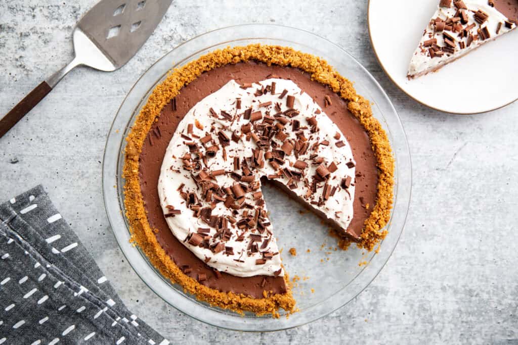 an overhead shot of a chocolate pie wit ha slice taken out. Whipped cream and chocolate shavings are spread across the top of the pie.