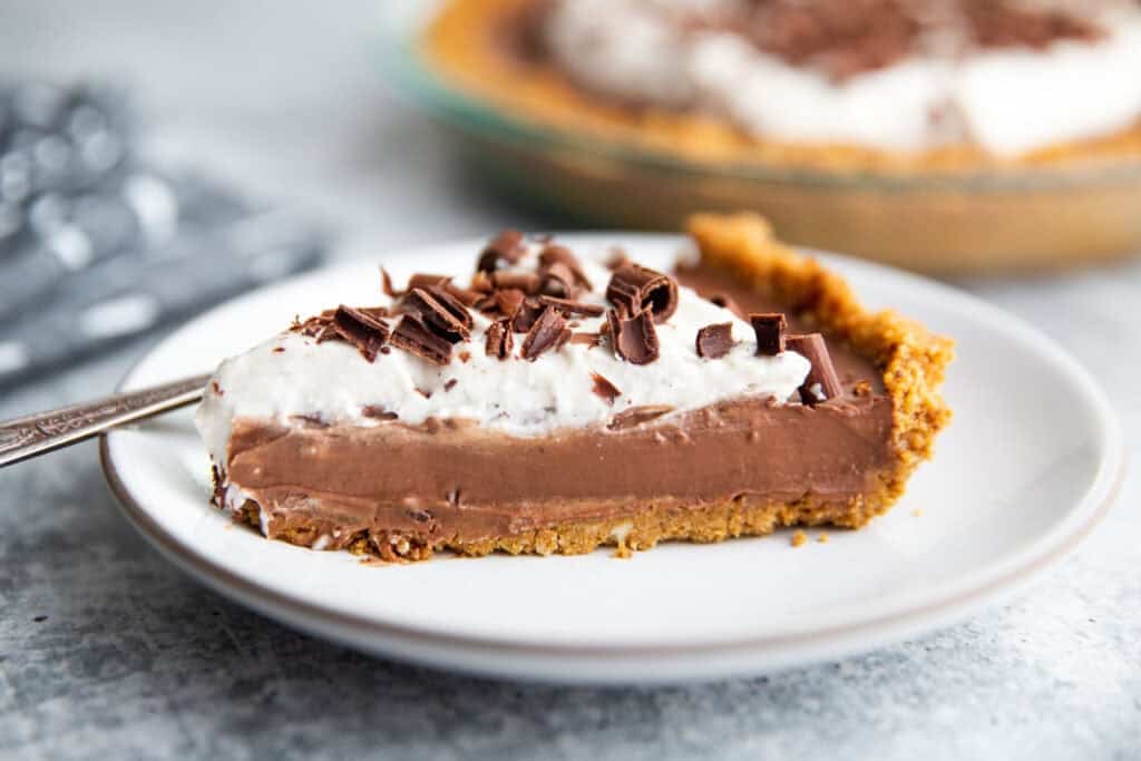 close up photo of a slice of pudding pie on a white plate topped with whipped cream and chocolate shavings