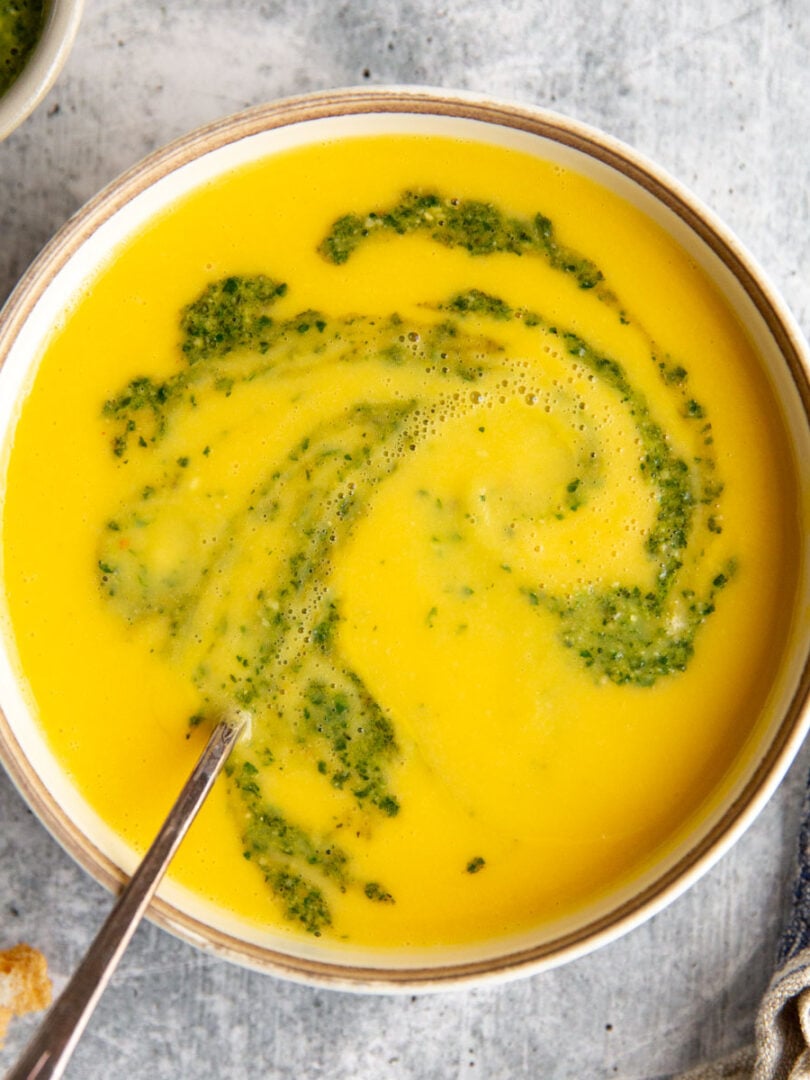 a bowl of cream yellow squash soup with a swirl of pesto on top.