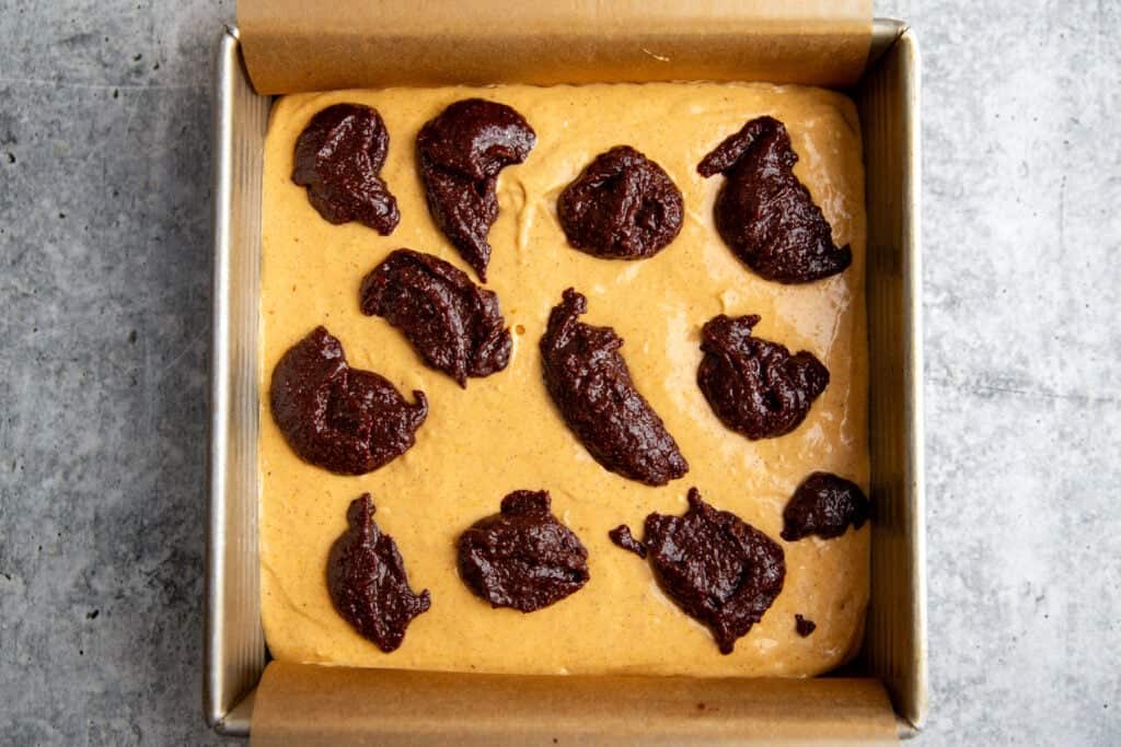 cheesecake filling atop the brownie batter in the brownie pan with drops of brownie batter on top waiting to be swirled