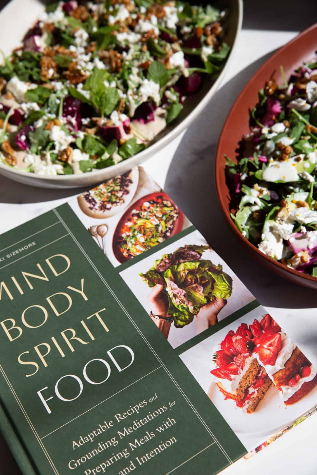 two roasted beet salads in serving trays finished with various toppings next to the cookbook Mind Body Spirit Food