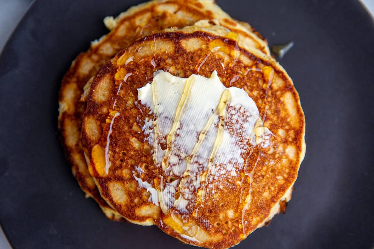 Sweet corn cakes stacked on top of each other on a black plate with butter spread on top of the pancake