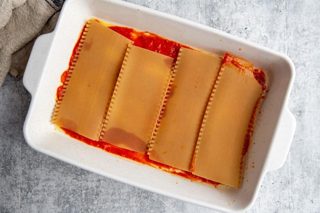 A white casserole dish lined with sauce and the first layer of lasagna noodles.