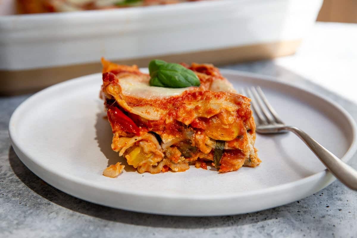 A slice of roasted vegetable lasagna made with gluten-free noodles on a white plate with a fork resting on the side.
